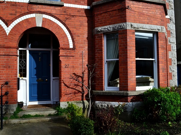 Exterior photograph of a period red brick facade home with white bay sash window, blue period front door, granite steps and window sills.