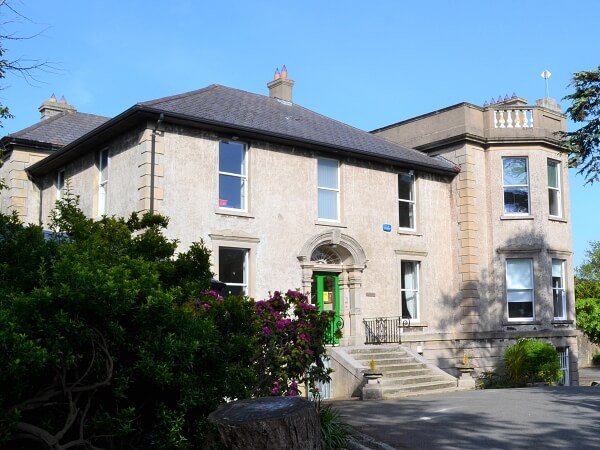 Photo at an angle of a large, estate period house, main building is square and side part of the building is protruding with bay windows. Granite steps up to a green door with a fan window above it. Sash window on the side and front are new bespoke sash windows by Bolger Sash windows. Large green bush and tree stump in the left foreground