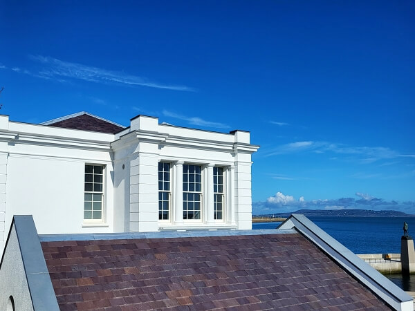 Side view of the top of Dun Laoghaire Baths showing new replica timber sash windows by Bolger Sash Windows. Part of the top of a slate roof in the foreground. The right background shows blue skies and Howth in the far distance