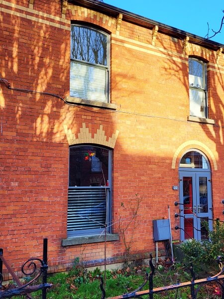 Side angle photo of a two storey, red brick, period house with sash windows restored and with BENGglas vacuum glass by Bolger Sash Windows. Downstairs window, door and meter box are painted sky blue, Upstairs frames are painted grey. Grass and small shrubs slightly in view in the front garden