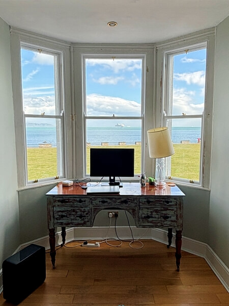 Desk with a computer screen and lamp on it, sitting in the alcove of a restored sash window by Bolger Sash Windows overooking a large green area, the sea, and a blue sky with white clouds